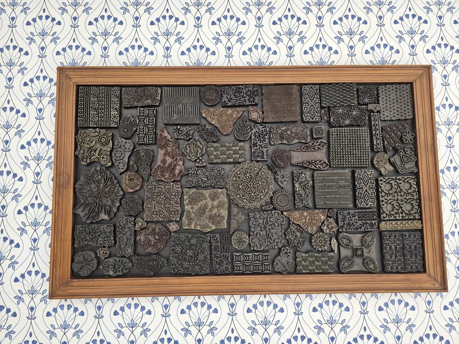 Carved printing blocks are arranged inside a wooden frame on patterned wallpaper.