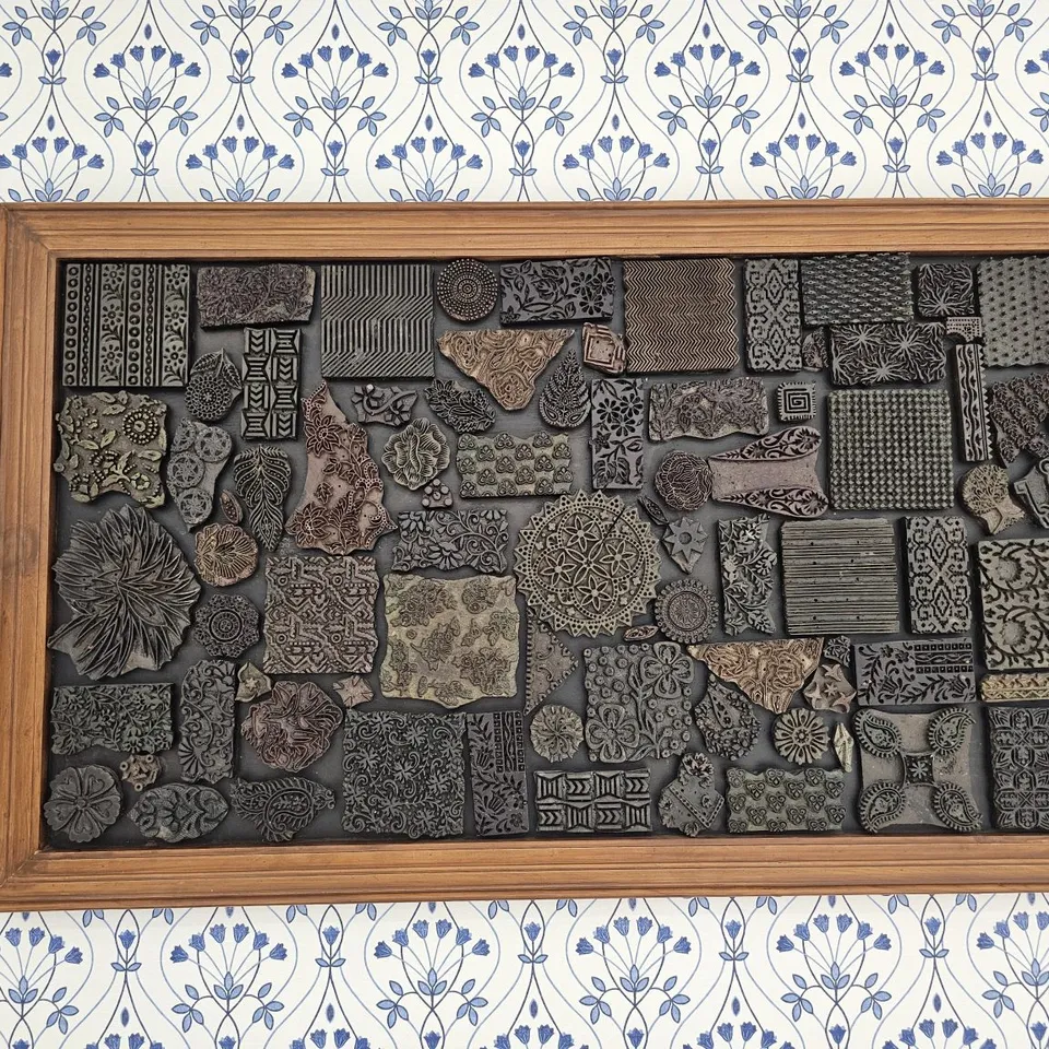 Carved printing blocks are arranged inside a wooden frame on patterned wallpaper.