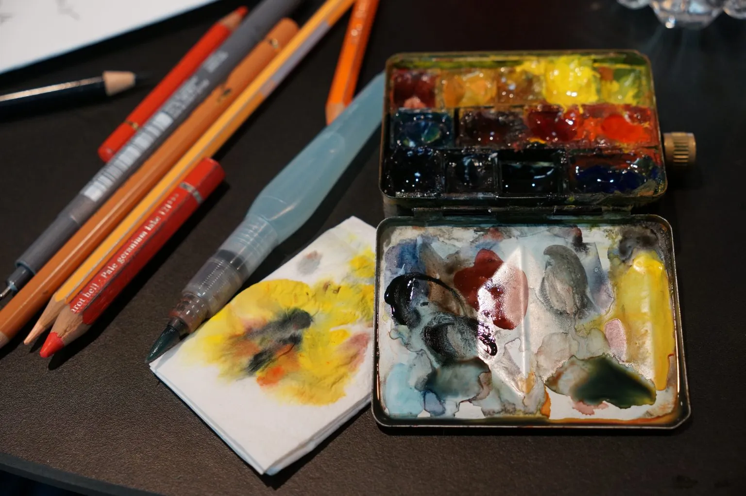 An open watercolor tin sits beside colored pencils, a water brush and stained paper.