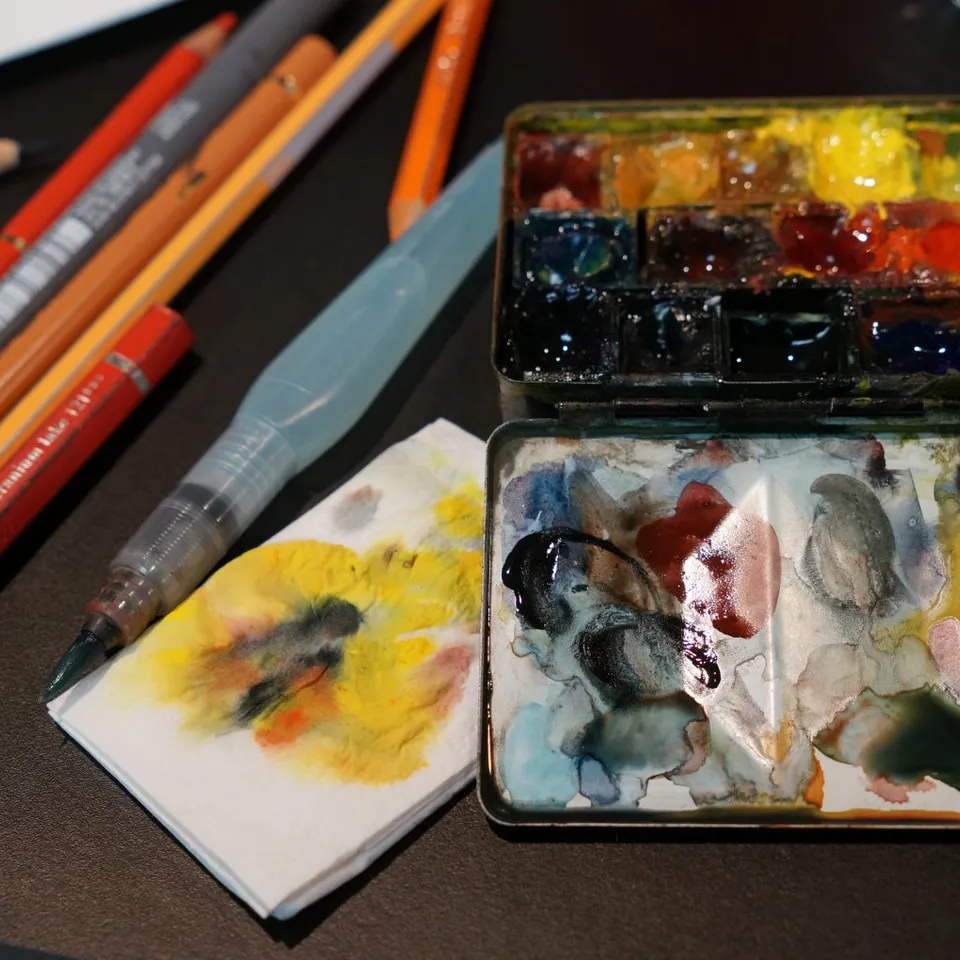 An open watercolor tin sits beside colored pencils, a water brush and stained paper.